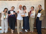 Six people standing together in front of a wall with large whiteboards covered in handwritten notes, each holding a small product and a certificate. The setting appears to be an indoor workshop or presentation space.