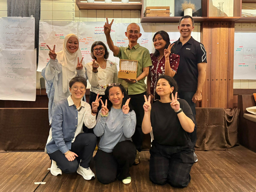 A group of nine people posing together in an indoor cafe or workshop environment with wooden shelves, whiteboards, and a hanging pendant light. Several individuals are making peace signs, and one person in the center holds a small box.