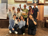 A group of nine people posing together in an indoor cafe or workshop environment with wooden shelves, whiteboards, and a hanging pendant light. Several individuals are making peace signs, and one person in the center holds a small box.